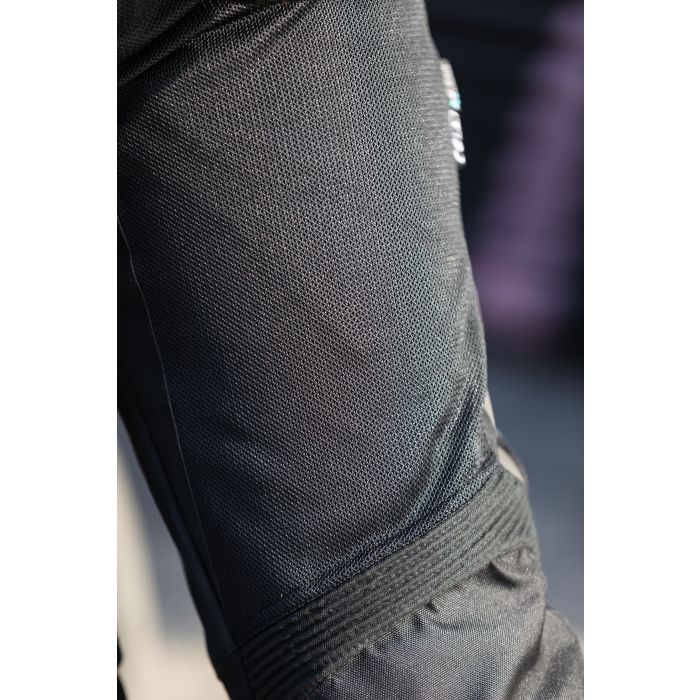 Primer plano de una tela negra del Pantalón Moto Verano Garibaldi Cool Typhoon. El material texturizado, similar a la malla, presenta detalles de costuras sobre un fondo difuminado con tonos oscuros y morados.